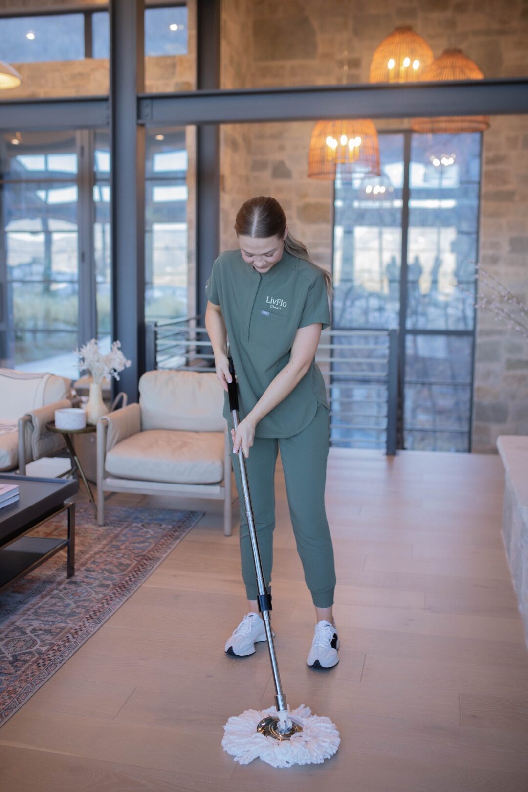 A LivFlo cleaner running a spin mop across a sunlit hardwood floor in a beautifully styled living room with leather chairs and woven pendant lights.