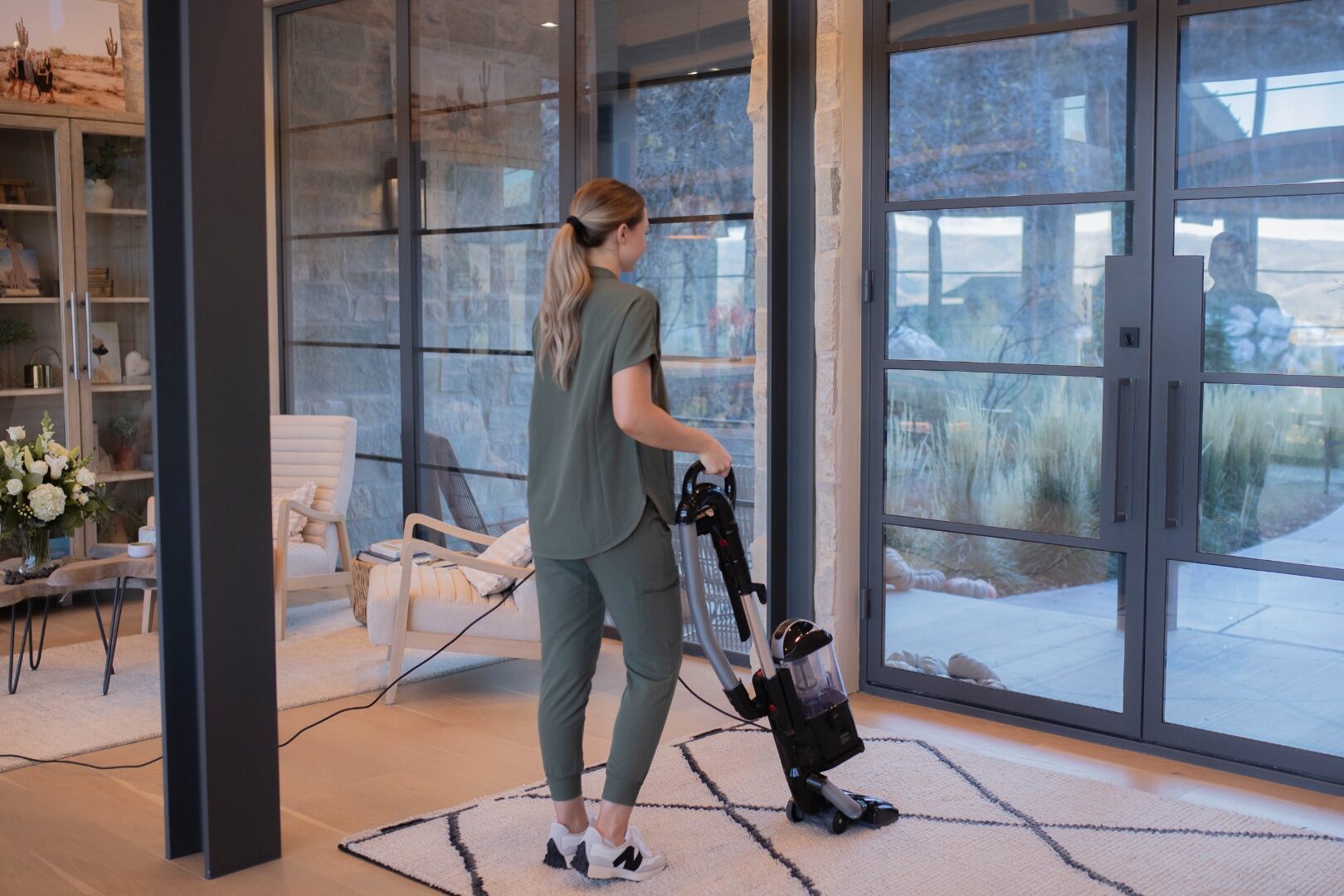 A LivFlo cleaner vacuuming the rug in a modern home with floor-to-ceiling glass doors opening onto a stone patio.