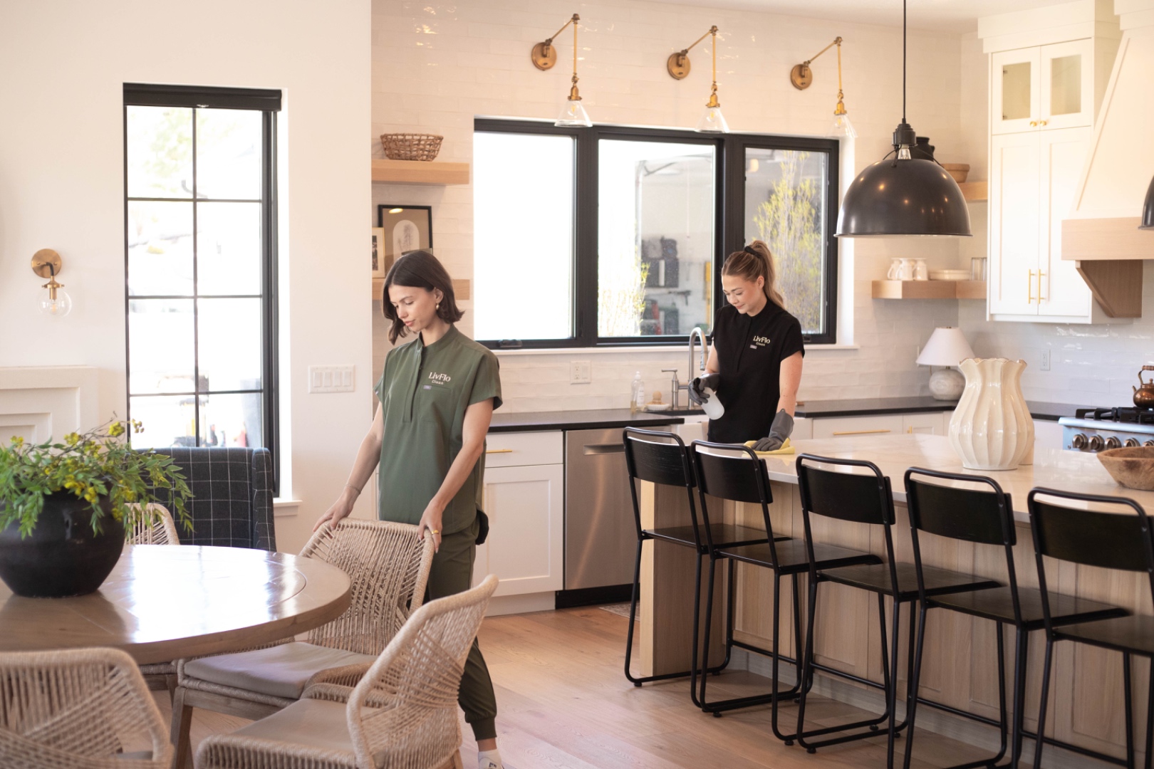 A LivFlo two-person team working in a bright, open kitchen and dining area — one tidying chairs, the other wiping the island.