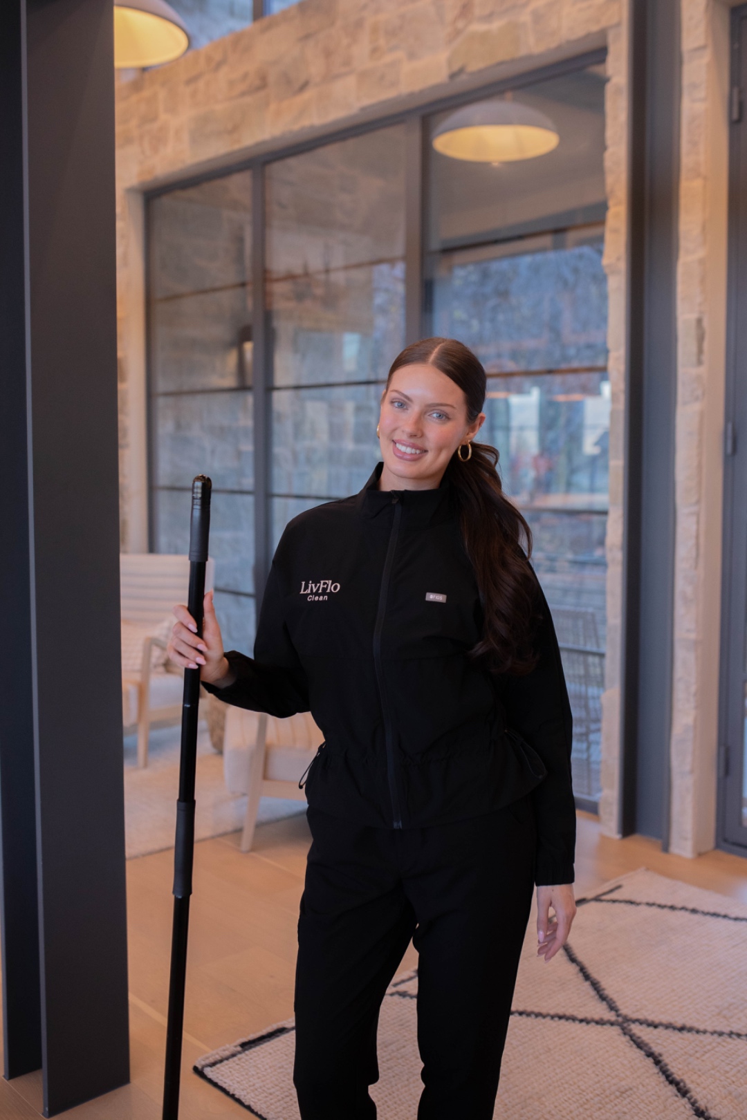 A LivFlo Clean team member in branded uniform holding a window-cleaning extension pole inside a modern stone-and-glass home.